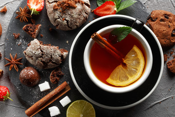 Fragrant tea in a black cup on a black plate with biscuits, lemon, cinnamon and fruits.