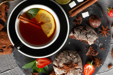 Fototapeta na wymiar Fragrant tea in a black cup on a black plate with biscuits, lemon, cinnamon and fruits.