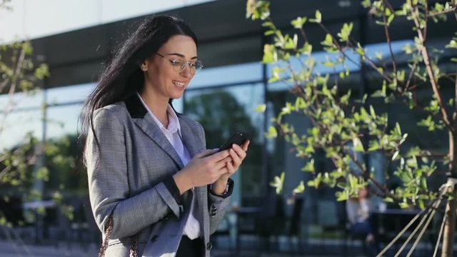 side view of smiling handsome professional woman walking on the street using smart phone mobile looking at display networking online business chat conversation internet searching sunshine sunny day