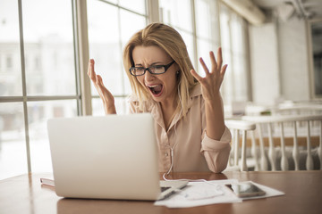 Young blonde businesswoman wearing eyeglasses and earphones in cafe sitting at table looking at laptop screen screaming angry shocked failed project