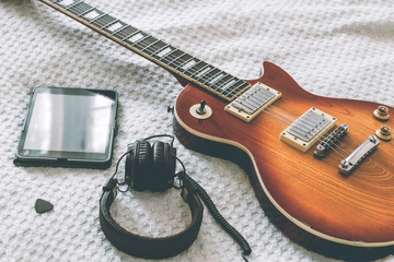 The electric guitar is on a white blanket next to the tablet and headphones