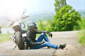Biker in outfit sits next to his motorcycle on the ground. Motorcyclist rests near the motorbike