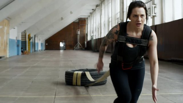 Tracking of determined young woman with tattoos pulling heavy tire and running in empty gymnasium