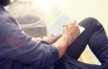 lifestyle, freelance, inspiration and people concept - close up of man with bag writing to notebook or diary sitting on city street bench