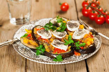 Baked eggplant with yellow and red bell pepper, tomatoes, garlic, goat cheese and chia seeds in plate on an old wooden rustic table