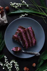 A piece of chocolate cake with blueberry cream on a gray plate. Dark background. Decor of flowers and hazelnuts. Flat lay