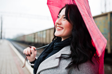 Brunette girl in gray coat with red umbrella in railway station.