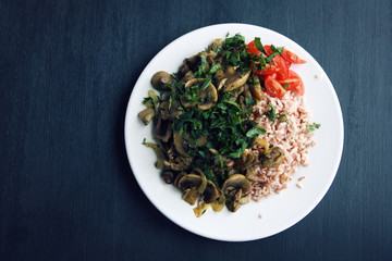 Red rice with mushrooms and cherry tomatoes. Vegan dish. European cuisine. Simple healthy lunch on the black background. Vegetarian rice dish on the white plate. Close up. Top view.