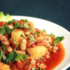 Vegetable stew with potatoes, white beans and tomatoes topped with parsley. European cuisine. Healthy vegan lunch on the white plate. Main course. Vegetarian dish. Toned square photo.