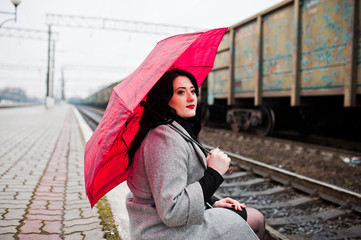 Brunette girl in gray coat with red umbrella in railway station.