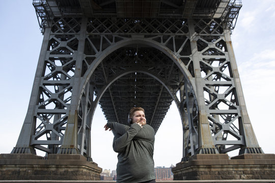 Overweight man stretching arms while standing below Williamsburg Bridge in city