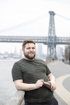 Portrait Of Smiling Overweight Man Holding Water Bottle While Standing Against Williamsburg Bridge In City