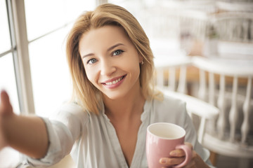 Young blonde woman in cafe sitting at table holding cup of hot coffee taking selfie pictures on smartphone looking camera smiling happy close-up