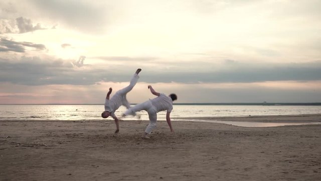 Two men in white clothes practicing capoeira on beach