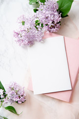 Top vew composition with feminine workspace mock up with empty card, lilac flowers and pink notebook on marble background