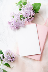 Top vew composition with feminine workspace mock up with empty card, lilac flowers and pink notebook on marble background