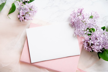 Top vew composition with feminine workspace mock up with empty card, lilac flowers and pink notebook on marble background