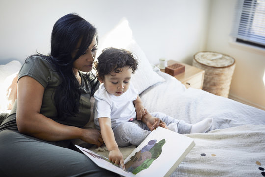 Cute son pointing at picture book while sitting by mother on bed