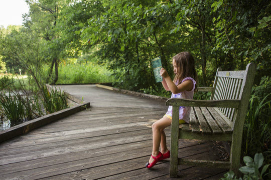 Girl Reading Book While Sitting On Bench At Park