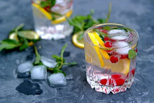 A cool drink, sparkling water with lemon, red currant, mint and ice in glasses on a dark gray concrete background