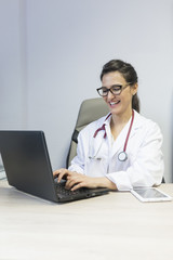 beautiful young doctor woman writing on laptop and smiling . She is wearing a white coat and a stethoscope. Medical concept.
