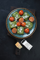 Turquoise plate with spinach ravioli, fried cherry tomatoes and grated parmesan, flatlay on a black stone background, vertical shot