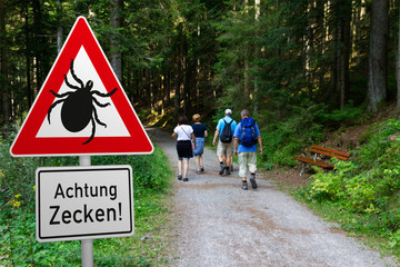 Schild mit Zeckenwarnung in Risikogebiet im Wald - mit Wanderern