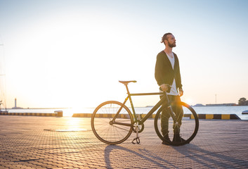 Young hipster man walking with bicycle during sunset or sunrise with sea port on background - obrazy, fototapety, plakaty