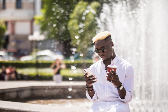 Portrait of handsome afro american young man listening music from phone drinking coffee to go on summer street in front of fountain