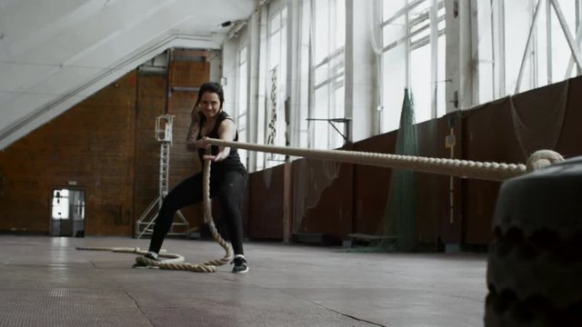 PAN of strong sportswoman in fitness clothes holding rope and pulling heavy tire towards herself during cross-training workout in empty gymnasium