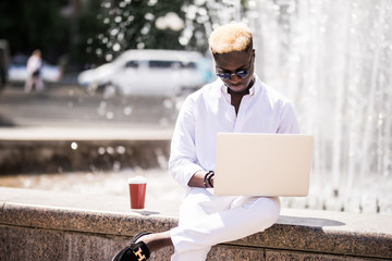 Young afro american man with the laptop sits at a fountain in summer street work outdoors