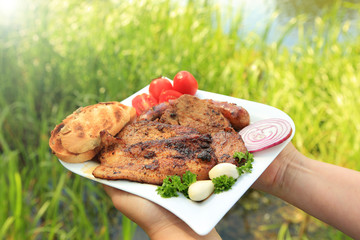 Kobieta trzyma w dłoniach grilowane mięso, pieczywo i warzywa na tle jeziora.