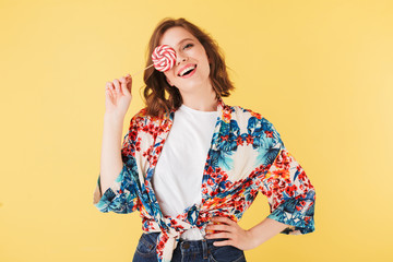 Portrait of pretty cheerful lady in colorful shirt standing and covering her face with lollipop candy while happily looking in camera on over pink background