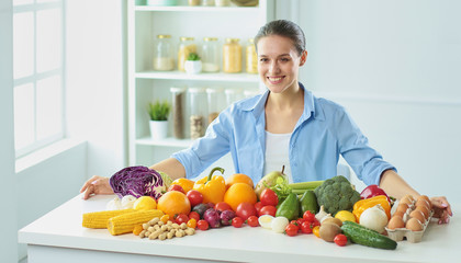 happy young housewife sitting in the kitchen preparing food from a pile of diverse fresh organic fruits and vegetables, selective focus