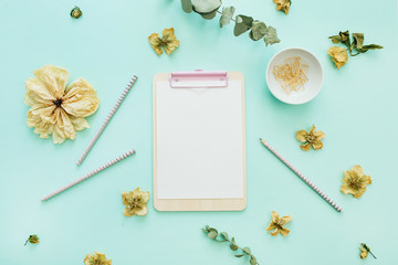 Flat lay home office desk workspace frame with space for text of clipboard, flowers and branches on blue background. Minimal blog hero header mock up with view from above.