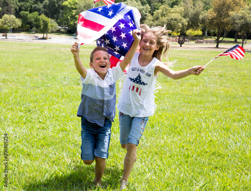 Adorable little girl and boy run on bright green grass holding american flag outdoors on beautiful summer day