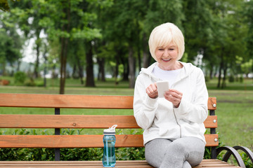 senior woman using smartphone while sitting on bench with sport bottle of water
