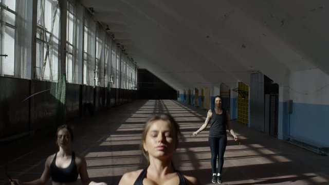 Tilt down of three sportswomen in fitness clothes jumping rope before camera while training in gym