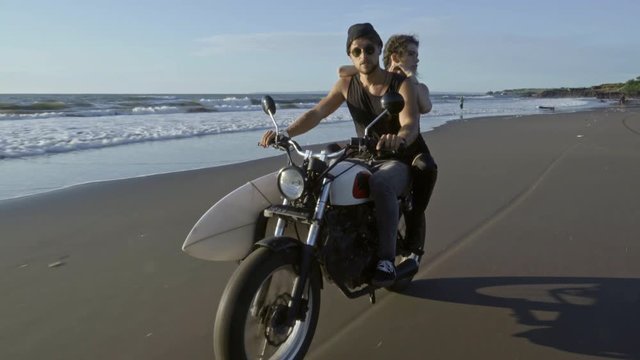 Couple of young surfers riding motorcycle on beach and enjoying nature, water splashing under motorbike wheels