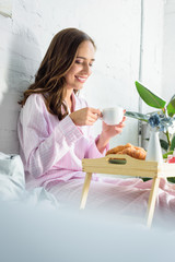 beautiful smiling woman in pink pajamas drinking coffee for breakfast in bed
