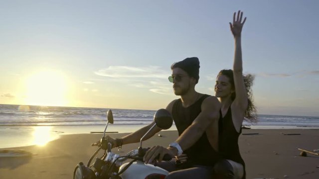 Young couple riding motorbike on the beach during sunset, woman making sign of the horns gesture and sticking out tongue to camera