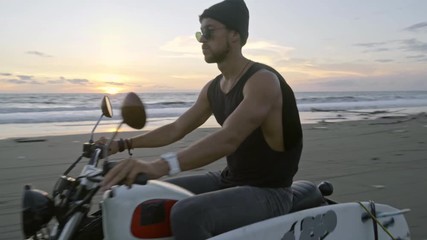 Young man beanie and sunglasses riding motorcycle with surfboard attached to it along ocean coast during sunset - Powered by Adobe