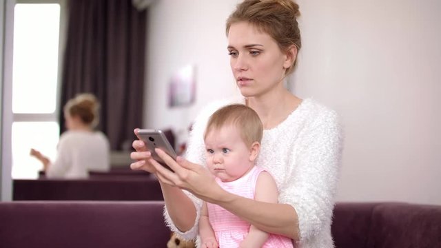 Woman with baby holding phone at home. Beautiful mother with baby looking smartphone. Modern mom with child holding mobile in hands. Working mother at home. Female everyday life