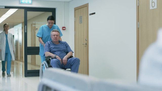 In the Hospital Hallway, Nurse Pushes Senior Man in the Wheelchair, Patients wait for their Doctor, Busy Professional Personnel Walking. Shot on RED EPIC-W 8K Helium Cinema Camera.