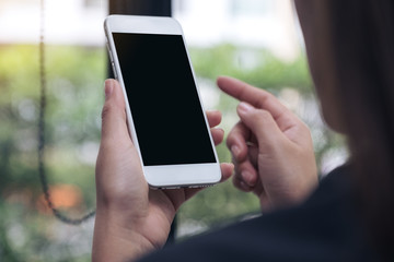 Mockup image of woman holding and pointing at a white mobile phone with blank black desktop screen in cafe