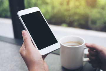 Mockup image of hands holding white mobile phone with blank black desktop screen while drinking coffee in cafe