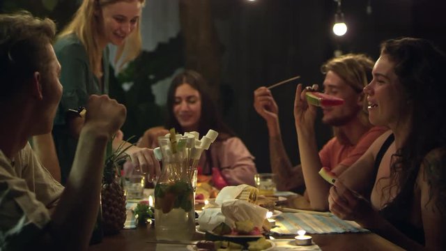 Group of young people sitting at table and enjoying tropical fruits, woman making fruit canapes