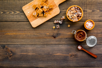 Cook caramel sauce. Melted caramel in glass jar, caramel cubes on cutting board, sugar and salt on dark wooden background top view copy space
