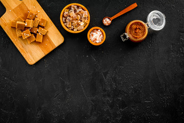 Salted caramel background. Melted caramel in glass jar, caramel cubes on cutting board, sugar and salt on black desk top view copy space