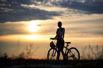 Fototapeta na wymiar Silhouette of sporty female cyclist, wearing sportswear, standing near her bicycle on trail. Slender incognito woman enjoying nature and observing wonderful landscapes and amazing sunset.